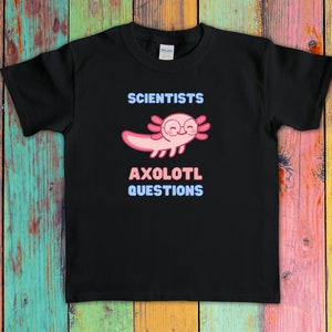 Puede incluir: Una camiseta negra de manga corta con un gr&aacute;fico de un ajolote rosa que lleva gafas redondas. El texto sobre el ajolote dice SCIENTISTS y el texto debajo dice AXOLOTL QUESTIONS en letras de color azul claro y rosa en la parte frontal.