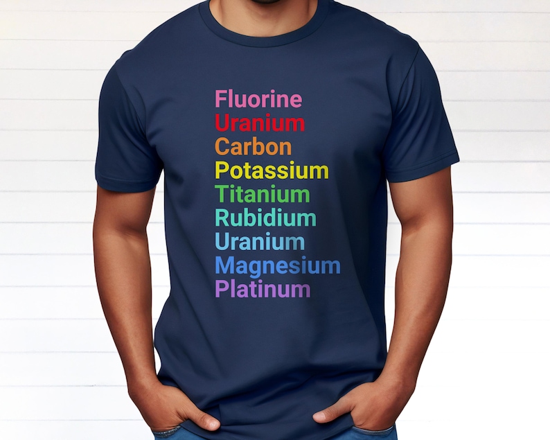 May include: A navy blue short-sleeved t-shirt featuring a vertical list of chemical elements printed in rainbow colors: Fluorine, Uranium, Carbon, Potassium, Titanium, Rubidium, Uranium, Magnesium, and Platinum, arranged to spell out a specific word.