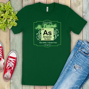 May include: Green t-shirt with a white graphic of a periodic table element. The element is arsenic, with the symbol As, atomic number 33, and atomic weight 74.922. The graphic also includes the text "100% All Natural", "Non-GMO", and "Gluten Free".