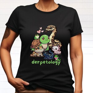 K&ouml;nnte beinhalten: Ein schwarzes Kurzarm-T-Shirt mit einer grafischen Illustration einer Schlange, einer Schildkr&ouml;te, eines Frosches, eines Axolotls, einer Eidechse und eines Salamanders mit drei kleinen rosa Herzen. Darunter steht der Text derpetology in gr&uuml;nen Buchstaben.