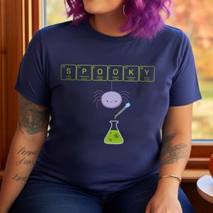 May include: Navy blue t-shirt with a Halloween-themed graphic. The word "SPOOKY" is spelled out using periodic table elements. A cartoon spider hangs above a beaker with green liquid. A fun and festive design.