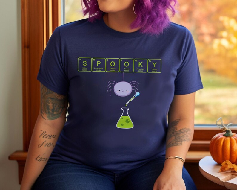 May include: Navy blue t-shirt with a Halloween-themed graphic. The word "SPOOKY" is spelled out using periodic table elements. A cartoon spider hangs above a beaker with green liquid. A fun and festive design.