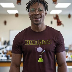 May include: A maroon t-shirt with the word "SPOOKY" spelled out using periodic table elements. Below the text is a cartoon spider, a beaker, and a pipette. The person in the image is wearing glasses and smiling.
