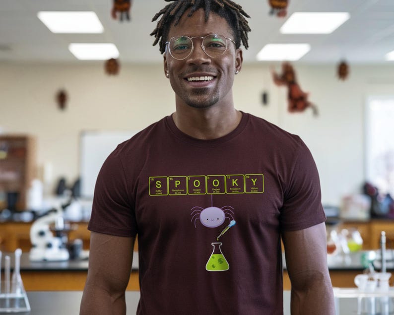 May include: A maroon t-shirt with the word "SPOOKY" spelled out using periodic table elements. Below the text is a cartoon spider, a beaker, and a pipette. The person in the image is wearing glasses and smiling.
