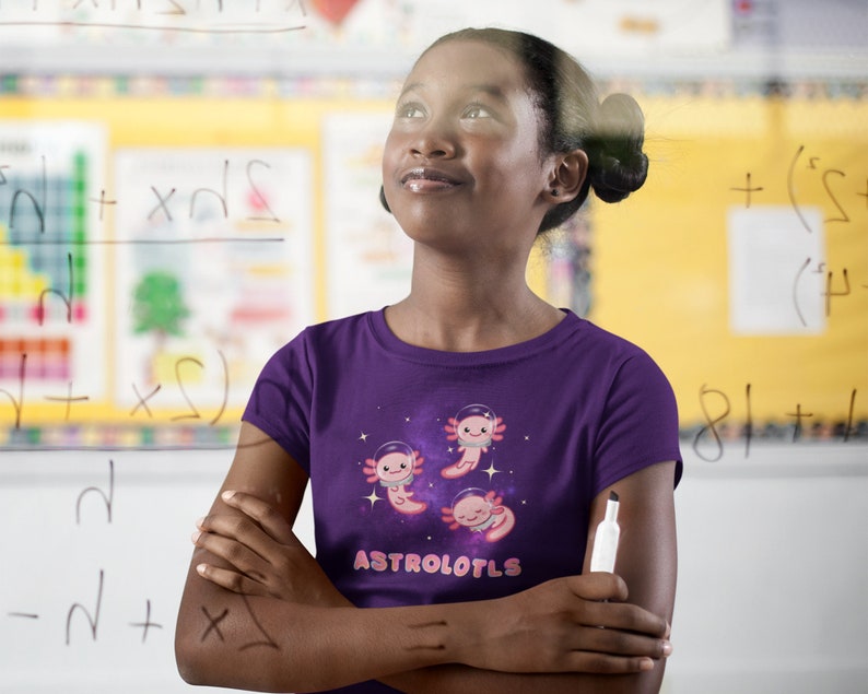 K&ouml;nnte beinhalten: Eine junge Person tr&auml;gt ein lila T-Shirt mit einem Weltraum-Motiv von Axolotln, die Astronautenhelme tragen. Der Text "ASTROLOTLS" ist auf das Shirt gedruckt. Die Person steht vor einer wei&szlig;en Tafel mit mathematischen Gleichungen darauf.