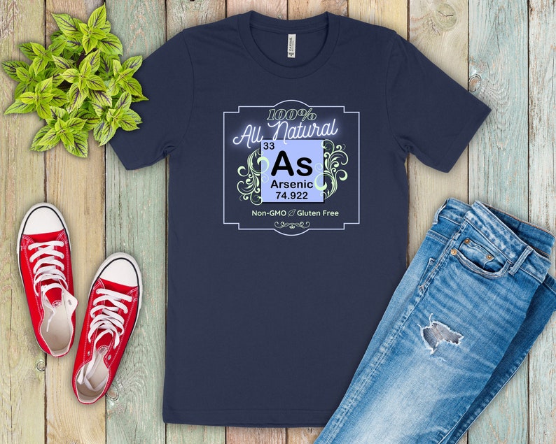 May include: A navy blue t-shirt with a white graphic of a periodic table element. The element is arsenic, with the symbol "As", atomic number 33, and atomic weight 74.922. The graphic is surrounded by a decorative border and the text "100% All Natural, Non-GMO, Gluten Free".