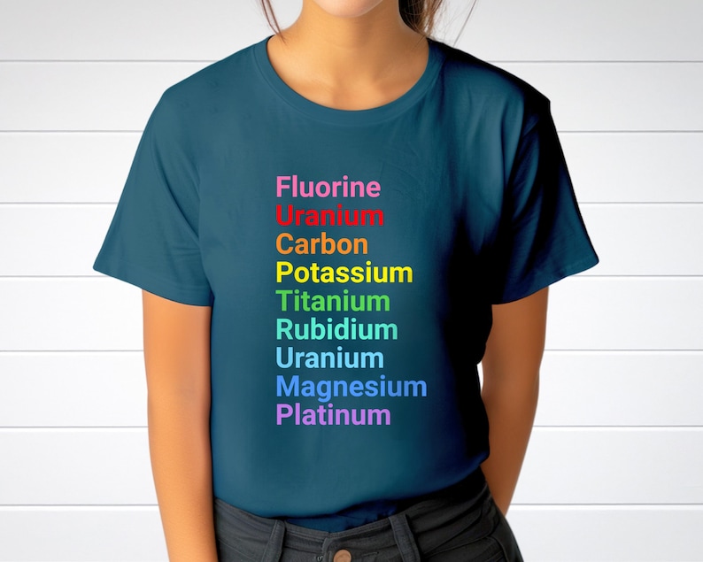 May include: A dark teal short-sleeved t-shirt featuring a vertical list of chemical elements printed in rainbow colors. The text reads: Fluorine, Uranium, Carbon, Potassium, Titanium, Rubidium, Uranium, Magnesium, and Platinum in descending order.