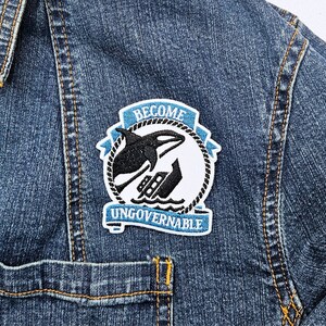 May include: A blue and white embroidered patch with a black orca jumping over a boat. The patch says "Become Ungovernable" in white text.