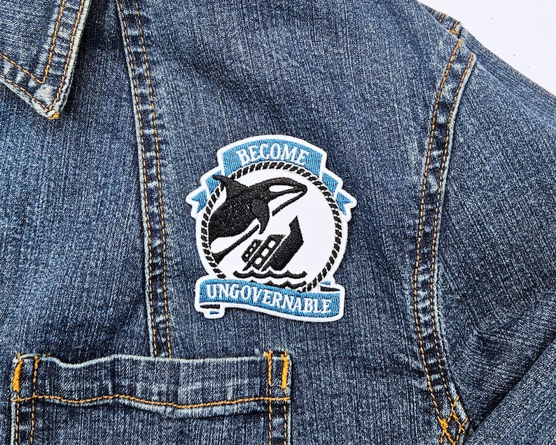 May include: A blue and white embroidered patch with a black orca jumping over a boat. The patch says "Become Ungovernable" in white text.
