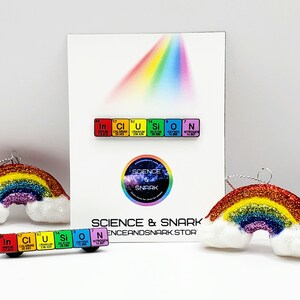 May include: Two rainbow-colored ornaments with a white cloud design. The ornaments are made of glitter and have a string for hanging. The ornaments are accompanied by two sets of colorful pins that spell out "Inclusion" and "Science & Snark".