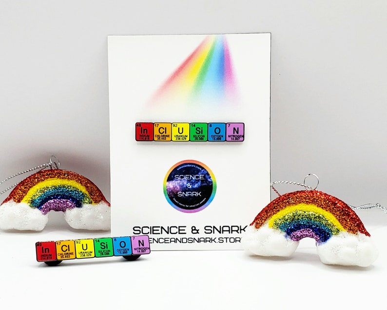 May include: Two rainbow-colored ornaments with a white cloud design. The ornaments are made of glitter and have a string for hanging. The ornaments are accompanied by two sets of colorful pins that spell out "Inclusion" and "Science & Snark".