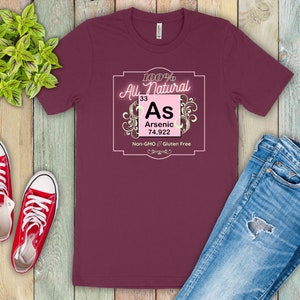 May include: A maroon t-shirt with a pink and white graphic design. The graphic features the chemical element arsenic, with the text "100% All Natural", "As", "Arsenic", "33", "74.922", "Non-GMO", and "Gluten Free".
