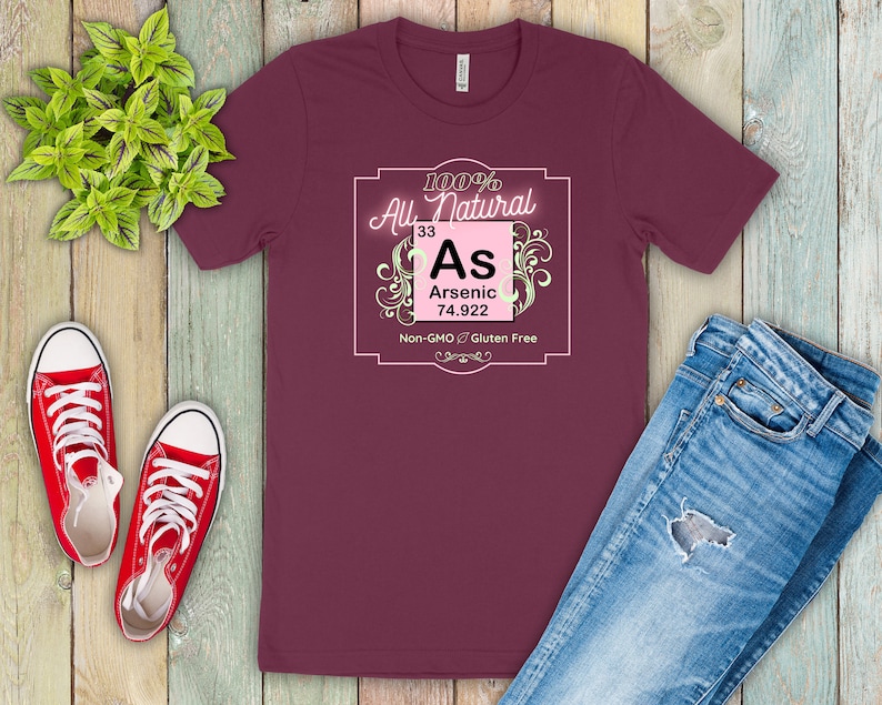 May include: A maroon t-shirt with a pink and white graphic design. The graphic features the chemical element arsenic, with the text "100% All Natural", "As", "Arsenic", "33", "74.922", "Non-GMO", and "Gluten Free".