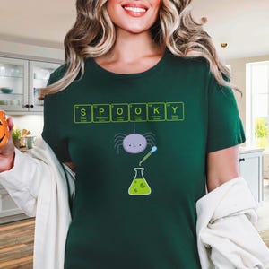 May include: Dark green t-shirt with the word "SPOOKY" spelled out using periodic table elements. A cartoon spider and beaker graphic are below the text. The person is holding an orange pumpkin-shaped mug.