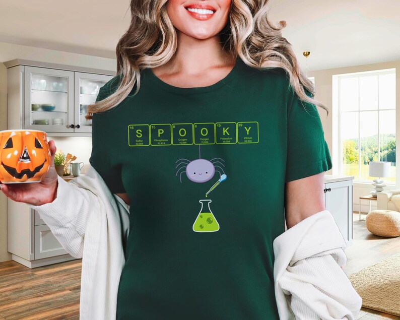 May include: Dark green t-shirt with the word "SPOOKY" spelled out using periodic table elements. A cartoon spider and beaker graphic are below the text. The person is holding an orange pumpkin-shaped mug.