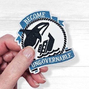 May include: Blue and white embroidered patch with a black orca silhouette jumping over a ship. The patch has a blue ribbon with the text "Become Ungovernable".