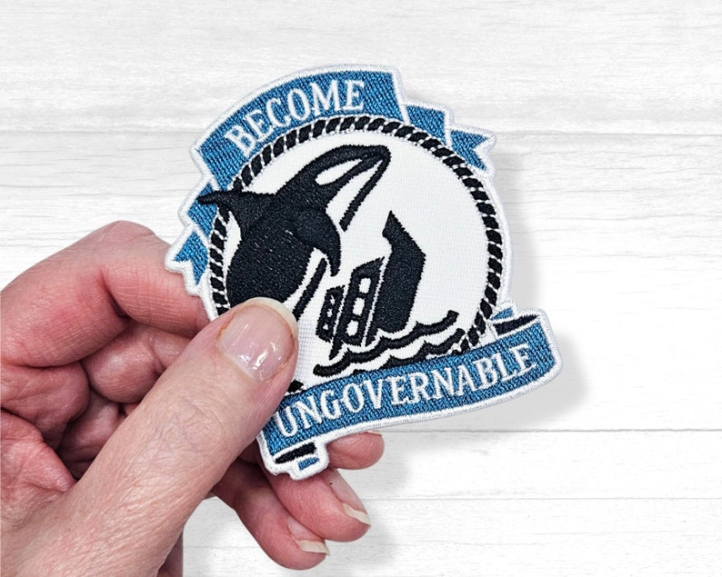 May include: Blue and white embroidered patch with a black orca silhouette jumping over a ship. The patch has a blue ribbon with the text "Become Ungovernable".