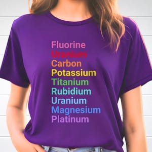 May include: A purple short-sleeved t-shirt featuring a vertical list of chemical elements printed in rainbow colors: Fluorine, Uranium, Carbon, Potassium, Titanium, Rubidium, Uranium, Magnesium, and Platinum, arranged in a column on the front.