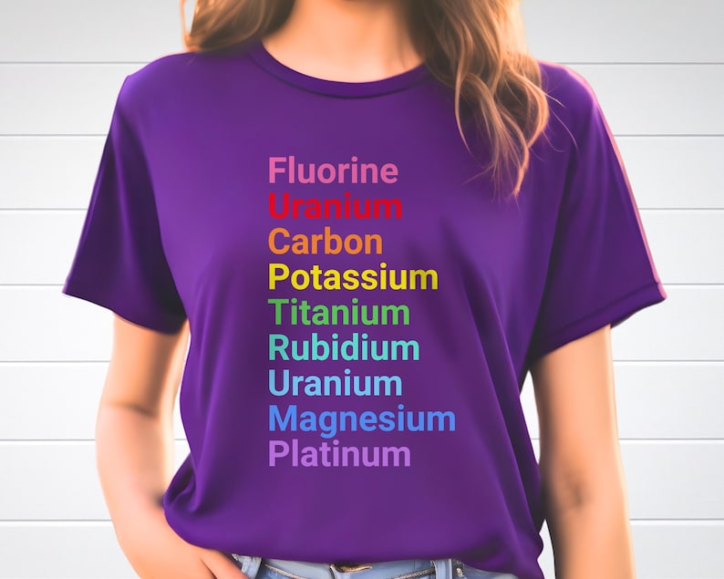 May include: A purple short-sleeved t-shirt featuring a vertical list of chemical elements printed in rainbow colors: Fluorine, Uranium, Carbon, Potassium, Titanium, Rubidium, Uranium, Magnesium, and Platinum, arranged in a column on the front.