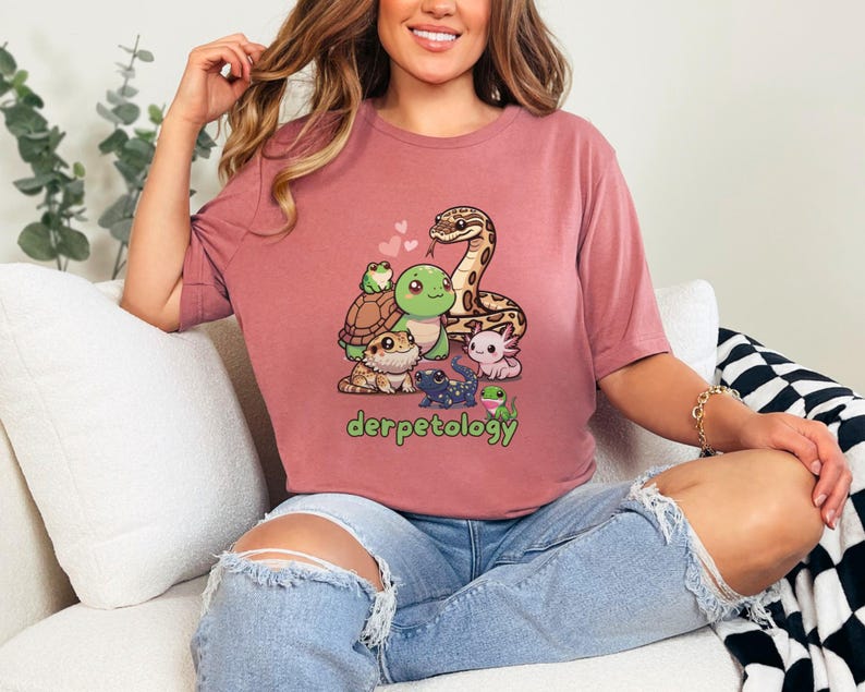 K&ouml;nnte beinhalten: Ein altrosa T-Shirt mit einer grafischen Illustration verschiedener Reptilien und Amphibien, darunter eine Schlange, eine Schildkr&ouml;te, ein Frosch und ein Axolotl, mit dem gr&uuml;nen Schriftzug "derpetology" unter der Tiergruppe und kleinen rosa Herzen.