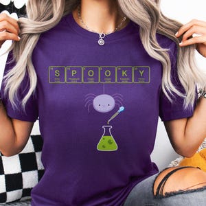 May include: A purple t-shirt with the word "SPOOKY" spelled out using elements from the periodic table. A cartoon spider and a green beaker are featured in the design. The shirt is made of a soft material.