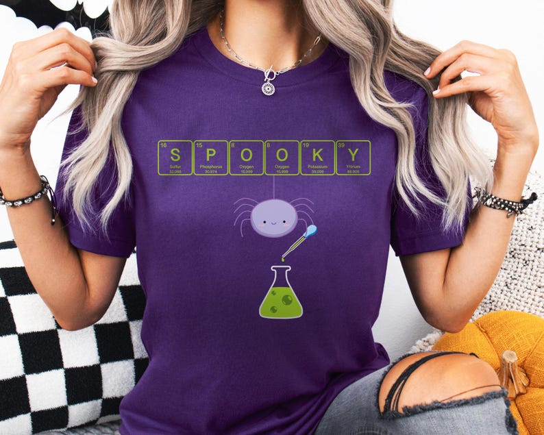 May include: A purple t-shirt with the word "SPOOKY" spelled out using elements from the periodic table. A cartoon spider and a green beaker are featured in the design. The shirt is made of a soft material.