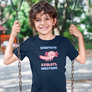 Puede incluir: Una camiseta de manga corta azul marino con un gr&aacute;fico de un ajolote rosa que lleva gafas redondas. El texto sobre el ajolote dice SCIENTISTS y el texto debajo dice AXOLOTL QUESTIONS en letras de bloque de color azul claro y rosa.