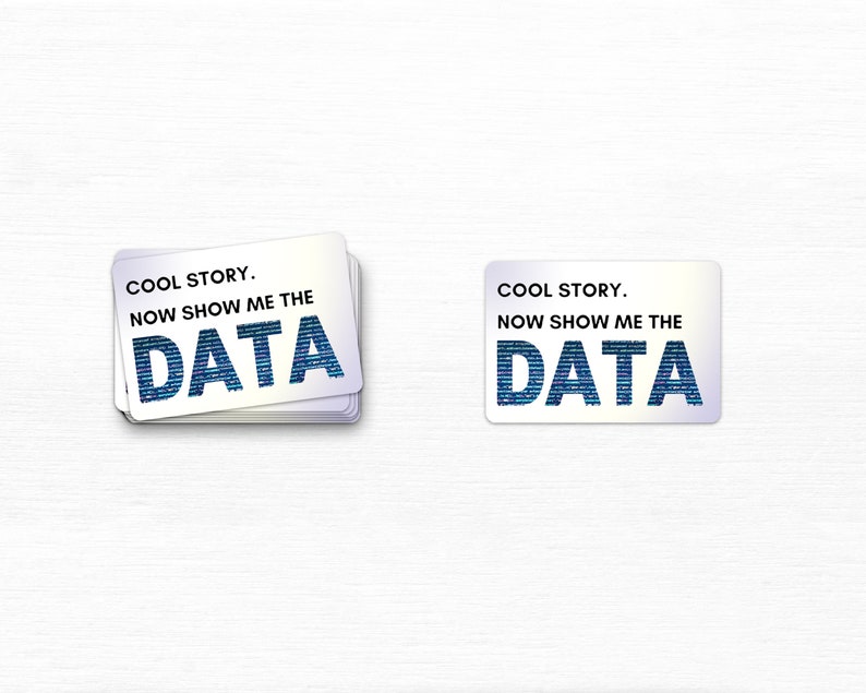 May include: A stack of white rectangular cards with rounded corners, each bearing the text "COOL STORY. NOW SHOW ME THE DATA" in a blue, code-like font. A single card is also shown. The background is a light wood grain.