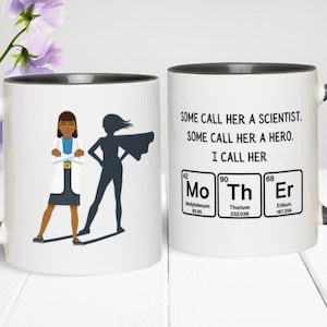 Two-tone ceramic mug featuring a cartoon scientist woman standing confidently with the shadow of a superhero behind her. The opposite side of the mug reads, “Some call her a scientist. Some call her a hero. I call her MoThEr,” with the word “Mother” spelled using periodic table elements (Molybdenum, Thorium, and Erbium). The mug is set on a white surface with purple flowers in the background.