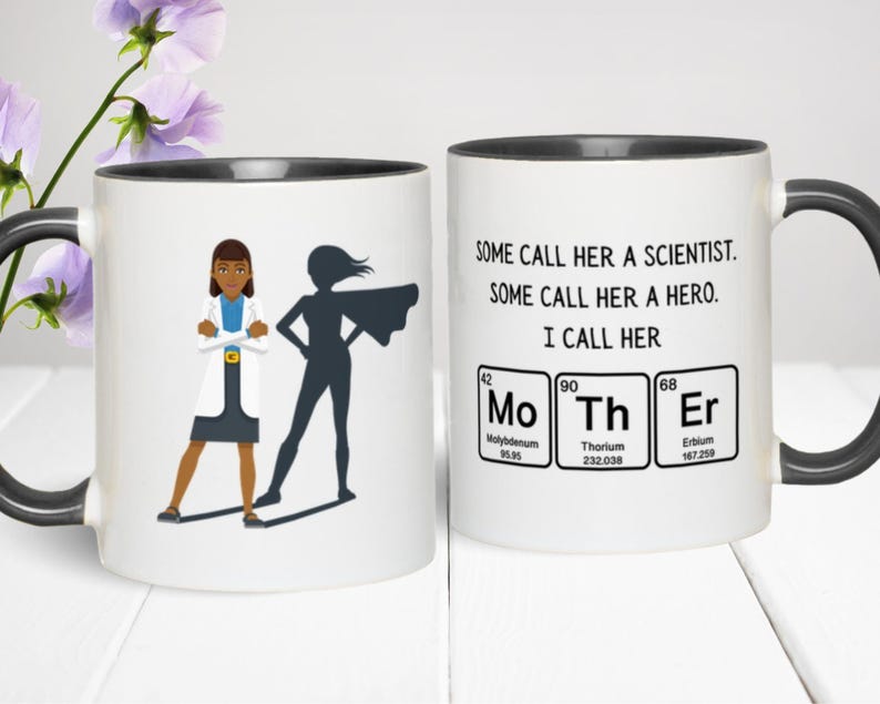 Two-tone ceramic mug featuring a cartoon scientist woman standing confidently with the shadow of a superhero behind her. The opposite side of the mug reads, “Some call her a scientist. Some call her a hero. I call her MoThEr,” with the word “Mother” spelled using periodic table elements (Molybdenum, Thorium, and Erbium). The mug is set on a white surface with purple flowers in the background.