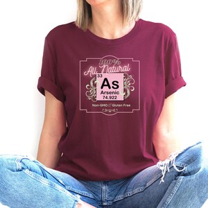 May include: A maroon t-shirt with a pink and white graphic of a periodic table element. The graphic says "100% All Natural 33 As Arsenic 74.922 Non-GMO Gluten Free".
