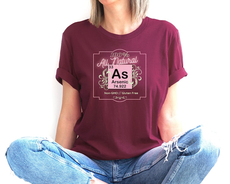 May include: A maroon t-shirt with a pink and white graphic of a periodic table element. The graphic says "100% All Natural 33 As Arsenic 74.922 Non-GMO Gluten Free".
