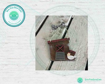 May include: Wooden farmhouse ornament featuring a brown barn with a red roof and a white chicken. The ornament has a black and white checkered bow and a string for hanging. The text "Sew Fun Sew Cute" is in a circle.