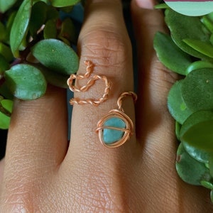 以下が含まれることがあります： ハート型のデザインとターコイズストーンが付いた、手作りの銅線リング。 リングは手の上に表示され、緑の葉に囲まれ、カラフルな円の中に「Orgonite Mami」というテキストが表示されています。