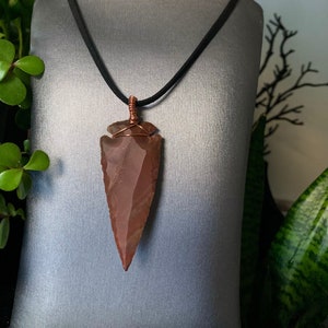 May include: A reddish-brown arrowhead pendant on a black cord. The arrowhead is wrapped with copper wire and hangs from a black cord. The pendant is displayed on a gray mannequin. The text "Orgonite Papi" is in a circle in the upper right corner.