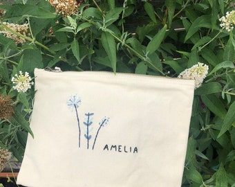 May include: A white canvas zippered pouch with a blue embroidered floral design and the name "Amelia" embroidered in black.