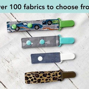 May include: Four fabric pacifier clips with plastic fasteners. The top strap features blue race cars, the second is gray with white arrows, the third is solid blue denim, and the bottom has a brown leopard print. Text reads: Over 100 fabrics to choose from!