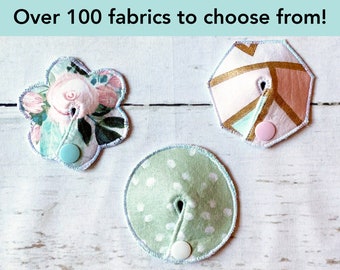 May include: Three fabric cord organisers with snap buttons. One is flower-shaped with a pink floral print and light blue snap, one is circular with a light green polka dot print and white snap, and one is hexagonal with a geometric print and pink snap.