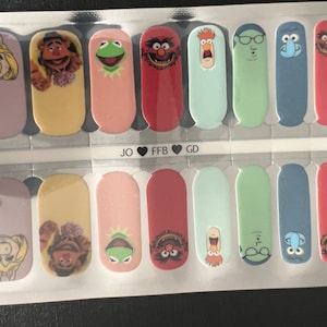 Könnte beinhalten: Ein Set mit 12 Nagelfolien mit Figuren aus den Muppets. Die Folien sind in zwei Reihen mit jeweils sechs Folien angeordnet, wobei jede Folie eine andere Figur zeigt. Zu den Figuren gehören Miss Piggy, Fozzie Bär, Kermit der Frosch, Animal, Gonzo und der schwedische Koch. Die Folien sind auf einem transparenten Hintergrund gedruckt und können direkt auf die Nägel aufgetragen werden.