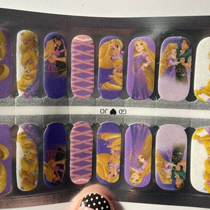 May include: A sheet of nail wraps featuring a purple background with images of Rapunzel and Flynn Rider from the Disney movie Tangled. The images include Rapunzel with long blonde hair, Flynn Rider with brown hair, and a purple and pink ribbon design.