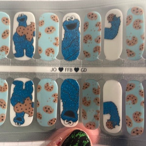 May include: Nail art strips with a light blue background, brown cookie designs, and images of a blue monster holding a cookie. The nail strips have the text "JO FFB GD".