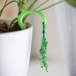 May include: A bright green plant pot hanger with a beaded tassel. The hanger is a curved, smooth plastic ring that fits over the edge of a white plant pot. The tassel features various shades of green beads.