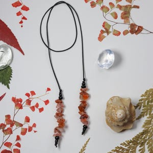 May include: A necklace with a black cord and two strands of orange-red gemstones. The gemstones are interspersed with small black beads. The necklace is displayed on a white surface with decorative elements including red leaves, a seashell, and clear stones.