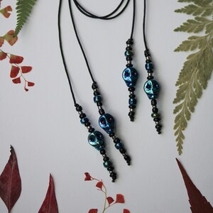 May include: A black cord necklace with three blue and black skull beads. The necklace is on a white background with green and red leaves.