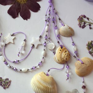 Könnte beinhalten: Eine handgefertigte Halskette mit Muscheln und Perlen in Lavendel-, Weiß- und Beigetönen. Die Halskette enthält Muscheln, sternförmige Perlen und Kristallakzente. Getrocknete Blumen sind um die Halskette herum angeordnet.