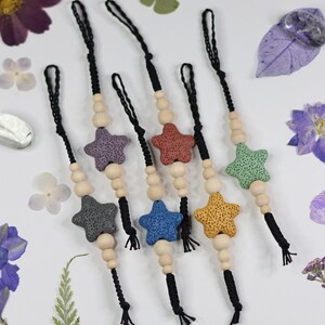 May include: A collection of six star-shaped ornaments in various colors, including gray, purple, red, blue, yellow, and green. Each ornament is strung on a black cord with wooden beads. The background features dried flowers and leaves.