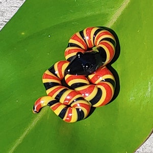 Könnte beinhalten: Eine gewundene Glasschlangenskulptur mit schwarzem Kopf und einem Körper, der mit roten, gelben und schwarzen Streifen versehen ist. Die Schlange liegt auf einem großen, leuchtend grünen Blatt auf einer Holzoberfläche.