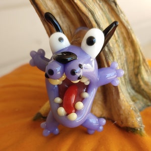 May include: A purple glass figurine of a cartoon dog with a wide open mouth and a red tongue sticking out. The dog has large white eyes with black pupils and black ears. The figurine is sitting on top of an orange pumpkin.