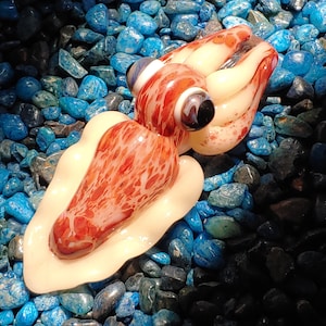 May include: A glass sculpture of a squid with red and white marbling. The squid is lying on a bed of blue and green pebbles.