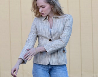 May include: A light beige blazer with a textured weave, featuring a notched lapel, two front pockets, and two brown buttons. The blazer is paired with light blue denim jeans.